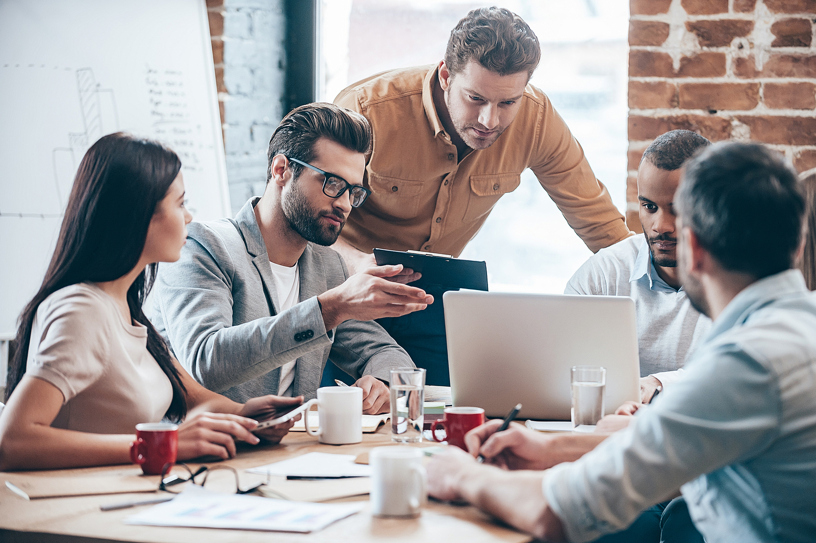 A diverse team of professionals collaborating around a laptop during a working meeting in a brick-walled office